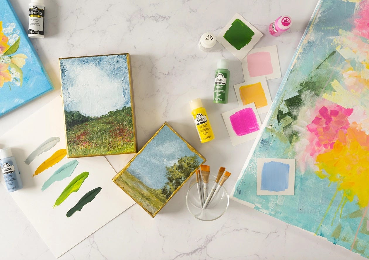 Art supplies arranged on a marble surface, including FolkArt acrylic paints, paintbrushes, color swatches, and landscape paintings.