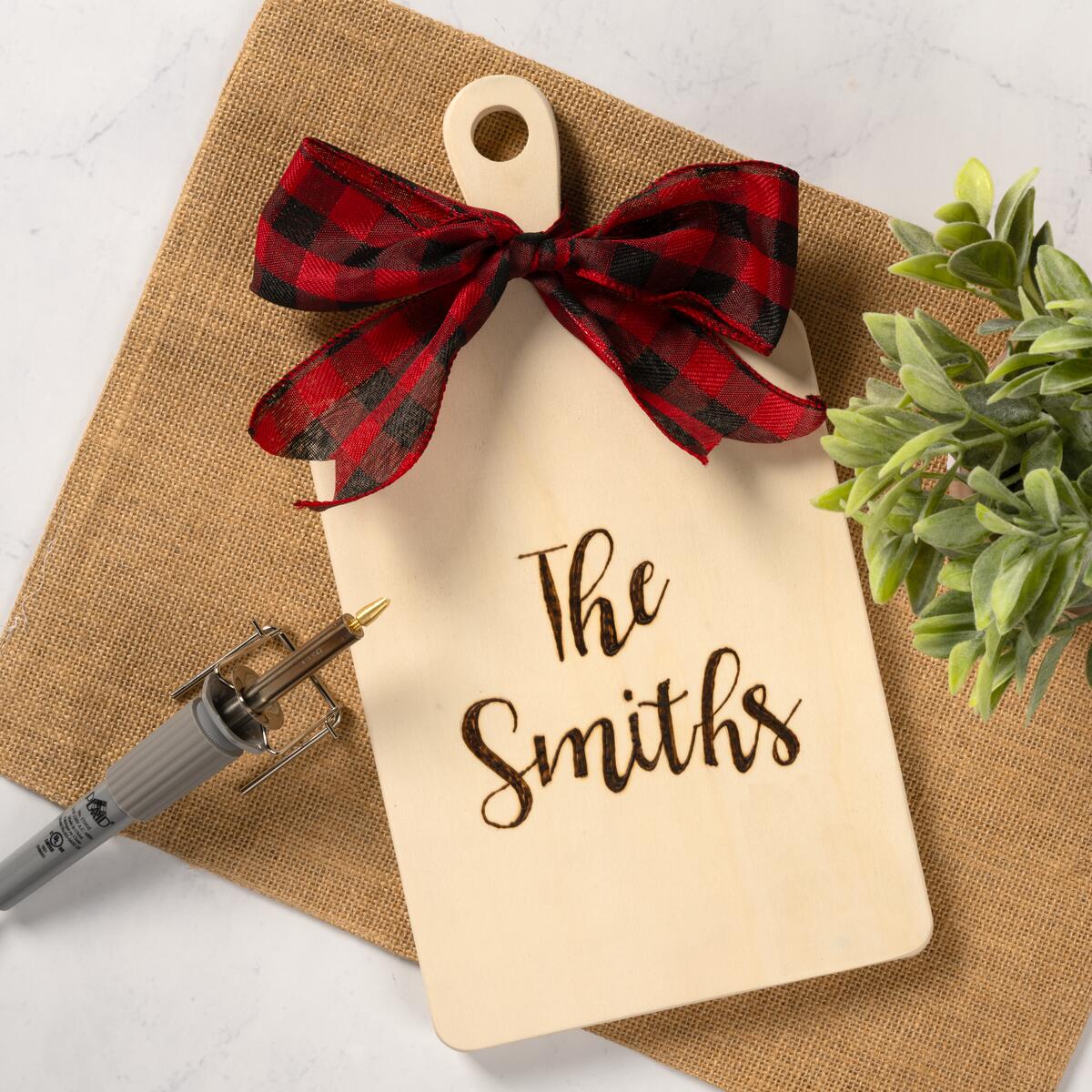 A decorative cutting board with a plaid ribbon and wood burned letters that say "The Smiths" 